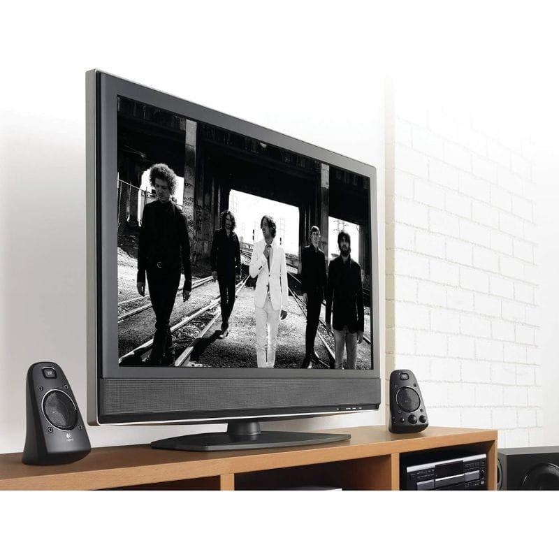 Logitech Z 623 2.1 Altavoces para PC con dos satélites negros con controles frontales, colocados junto a un TV en mueble de madera.
