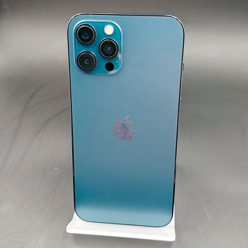 iPhone 12 Pro Max azul pacífico, traseira fosca com logo Apple, três câmeras grandes com flash, bordas retas em metal, suporte branco discreto.