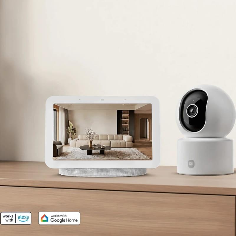 Xiaomi Smart Camera C302 3MP 2K WiFi 6 en blanco, cuerpo esférico con lente negra y base giratoria; junto a pantalla inteligente en mueble minimalista