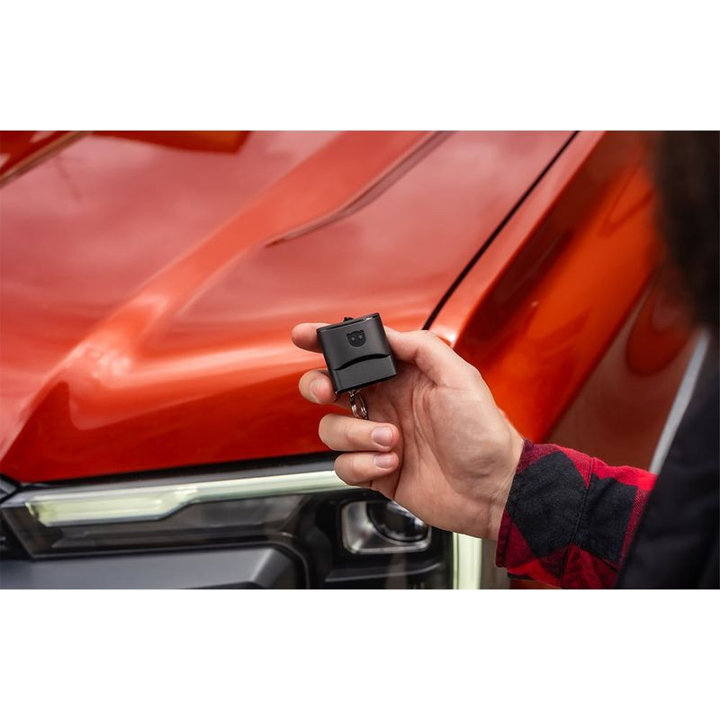 Scanner de diagnostic OBDeleven 3 compact noir, prise OBD rectangulaire avec logo, tenu en main près d’un capot orange et d’un phare LED de voiture.