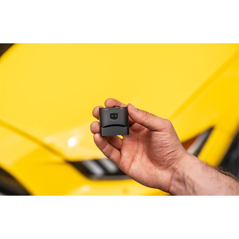 Petit adaptateur OBD noir tenu en main devant une voiture jaune; design compact, prise OBD intégrée, LED et logo visibles. Scanner de diagnostic OBDeleven 3