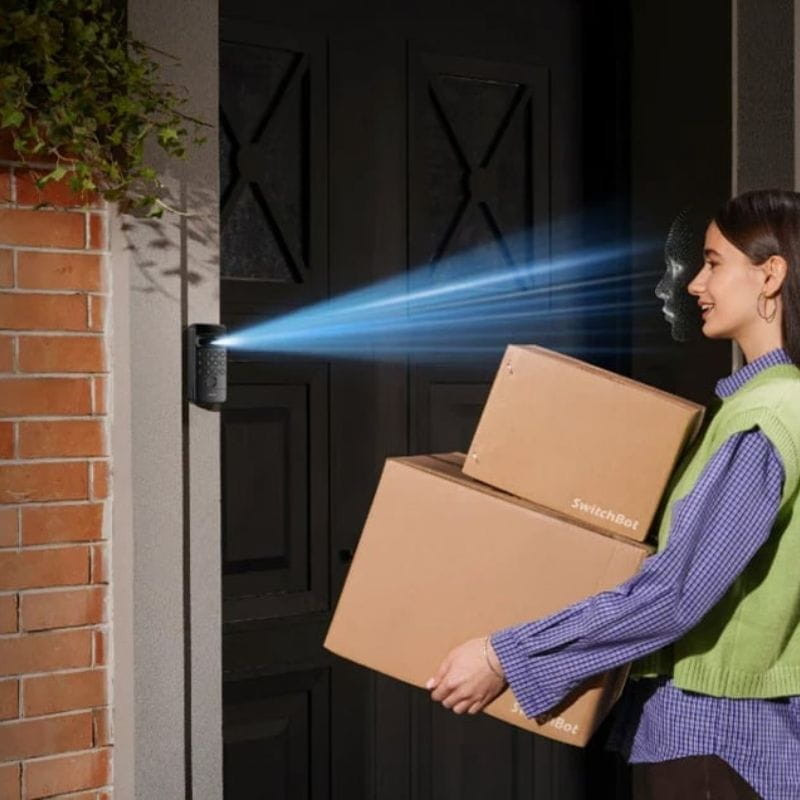 Teclado con reconocimiento facial SwitchBot Vision Lock instalado en puerta negra, cámara frontal proyecta haz azul, detecta rostro y permite acceso manos