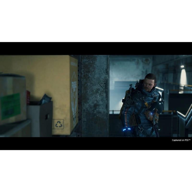 Death Stranding Directors Cut PS5: scène in-game sombre, héros en armure futuriste avec éclairages bleus, entrepôt industriel, carton recyclage visible à