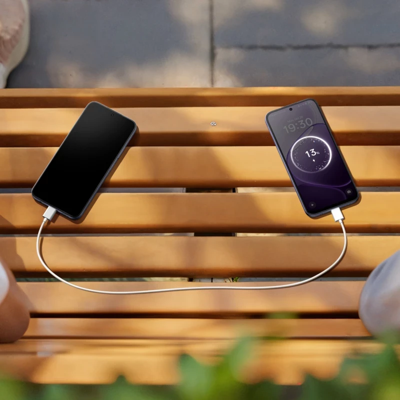 Cabo USB conectando dois smartphones sobre banco de madeira; tela da direita mostra carregamento a 13 por cento com relógio 19:30 e fundo lilás
