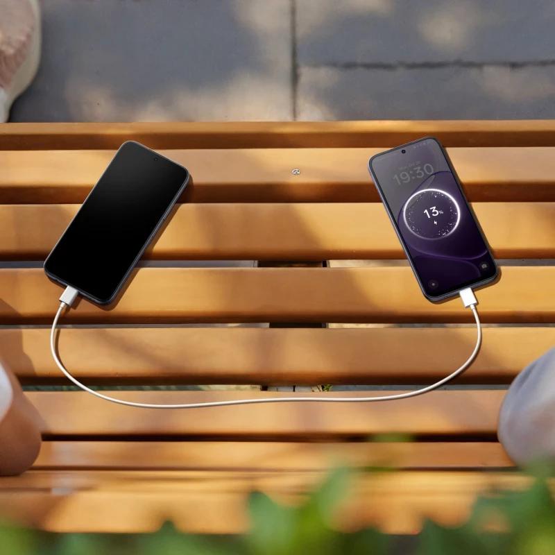 Cabo USB conectando dois smartphones sobre banco de madeira; tela da direita mostra carregamento a 13 por cento com relógio 19:30 e fundo lilás