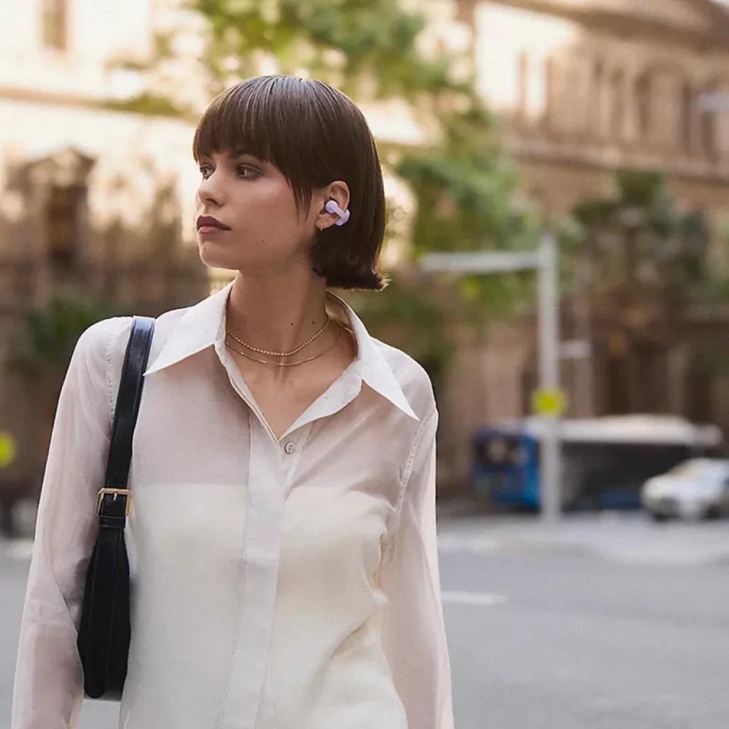 Auricular open-ear bege com clip preso à orelha; design leve e discreto, formato em arco, encaixe seguro e visível em uso urbano ao ar livre
