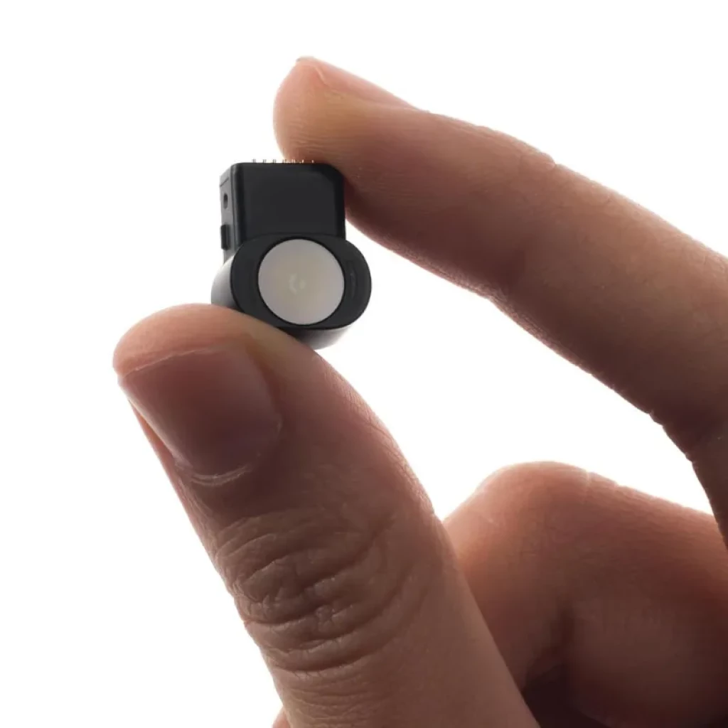 Módulo de cámara XREAL Eye negro con lente circular blanca, sujeto entre dedos para mostrar su tamaño pequeño y diseño minimalista
