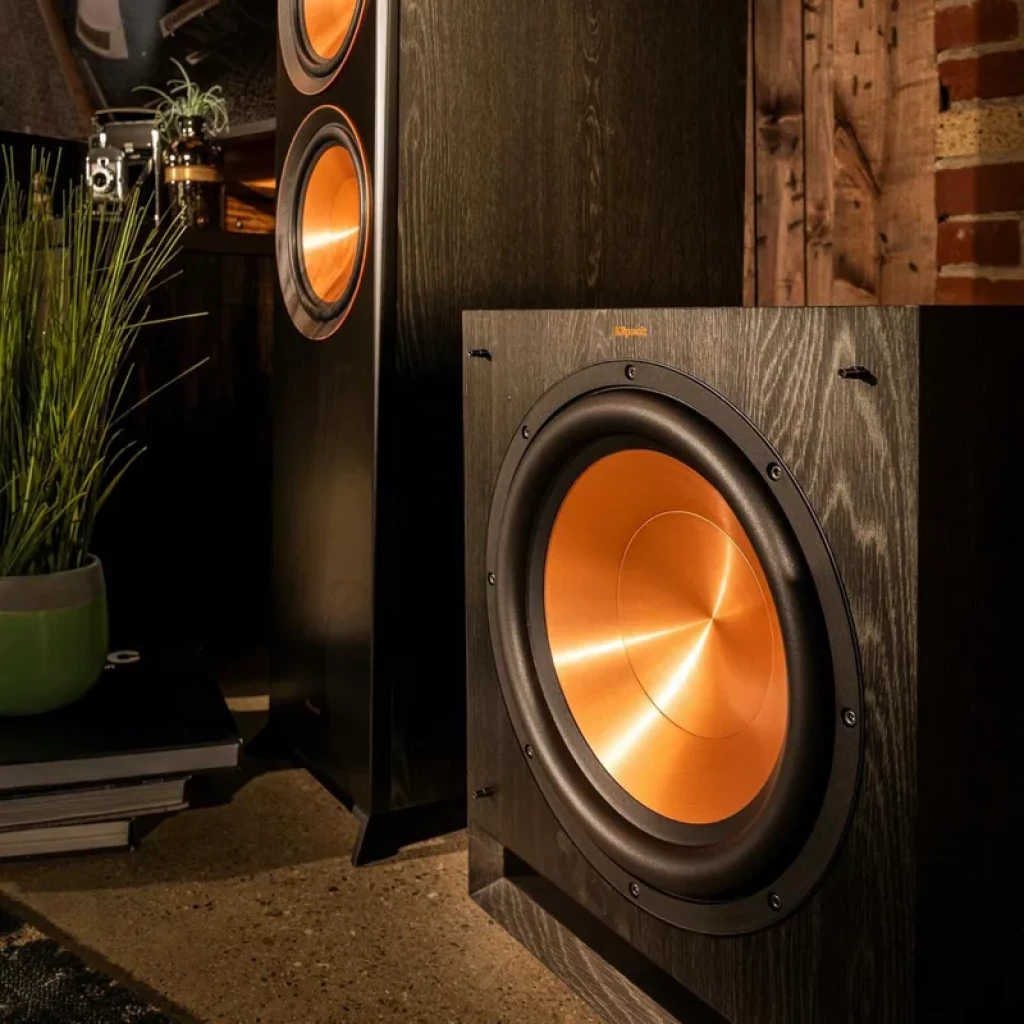 Klipsch SPL 120 CE de 12 pol com cone cobre, acabamento em madeira escura e borda emborrachada; subwoofer ativo com design robusto e moderno