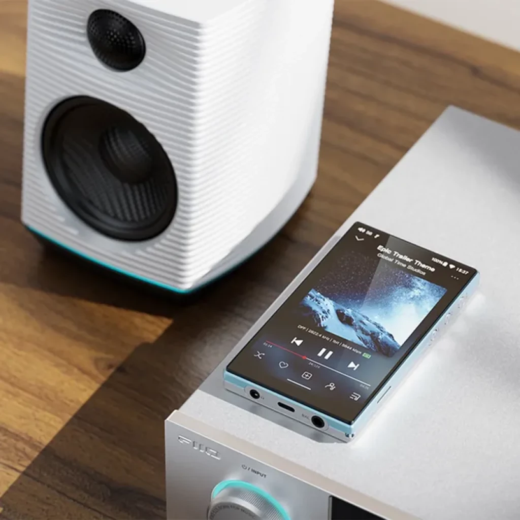 FiiO JM21 4+64 bleu posé sur ampli, écran affichant lecture musicale; double prise casque, boutons latéraux, design métal brossé près dune enceinte