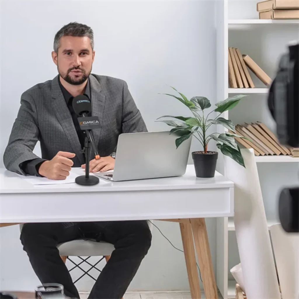 Comica HR-WM transmisor UHF de mano con cápsula supercardioide, rejilla metálica y cuerpo negro, montado en base de escritorio frente a presentador en set