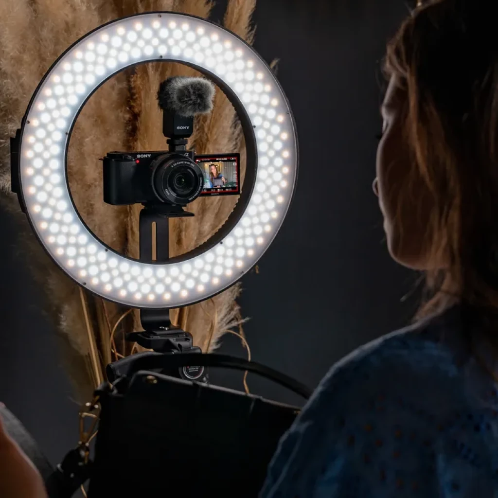 Cámara Sony ZV-E10 con lente 16-50mm montada en trípode, anillo de luz LED iluminando, micrófono con paraviento y pantalla abatible mostrando la grabación