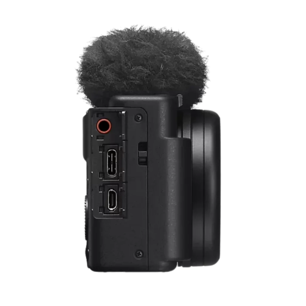 Lateral de la Sony ZV-1 II con micrófono peludo antiviento, puertos 3.5 mm, micro HDMI y USB, cuerpo compacto negro y lente 18-50 mm sobresaliente
