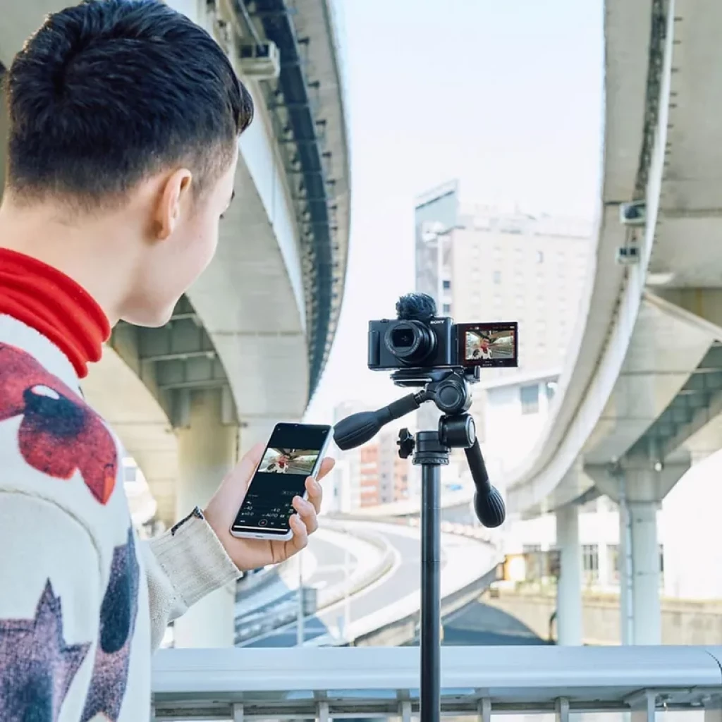 Sony ZV-1 II negra en trípode con mini empuñadura y micrófono peludo, pantalla abatible mostrando vlog; entorno urbano bajo pasos elevados, uso con