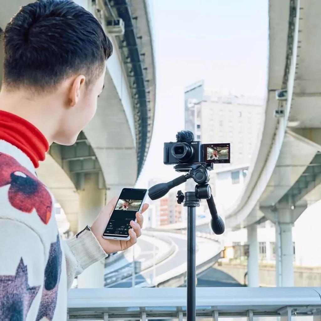 Sony ZV-1 II negra en trípode con mini empuñadura y micrófono peludo, pantalla abatible mostrando vlog; entorno urbano bajo pasos elevados, uso con