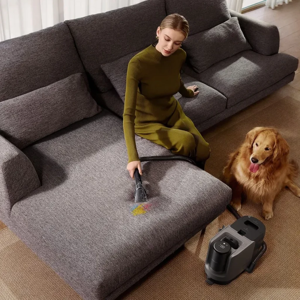 Dreame H15 Mix limpiando sofá con cepillo manual; elimina migas y manchas. Estación gris con depósito doble. Perro dorado observando junto a sofá gris