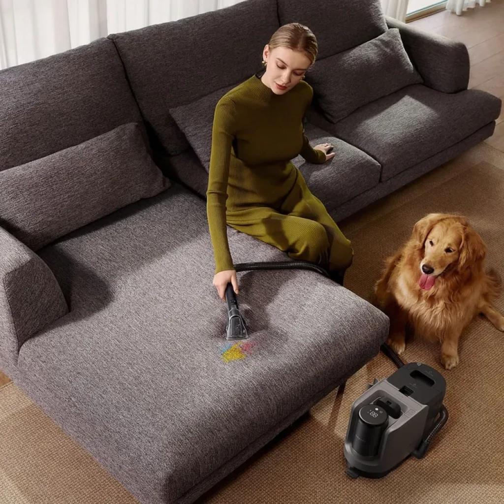 Dreame H15 Mix limpiando sofá con cepillo manual; elimina migas y manchas. Estación gris con depósito doble. Perro dorado observando junto a sofá gris