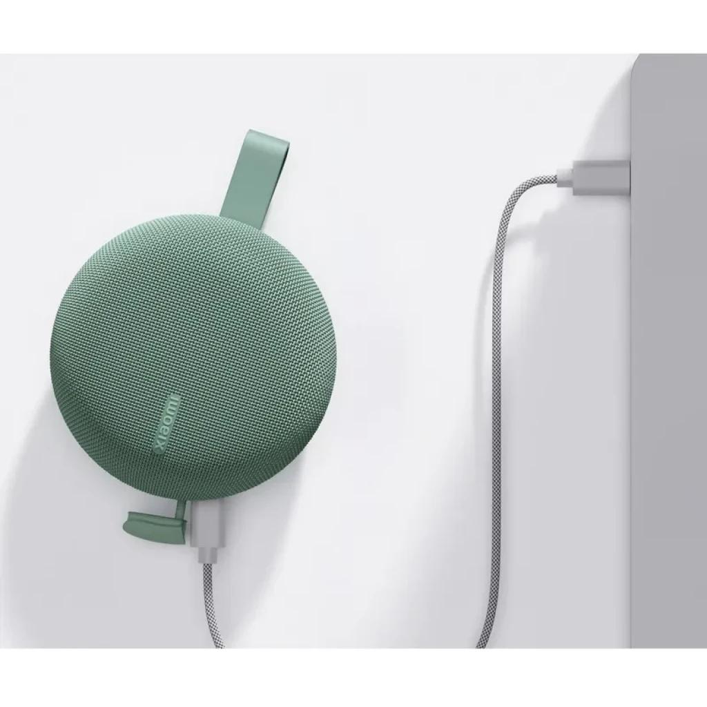 Altavoz portátil redondo verde con correa de silicona y rejilla textil, cargando por cable trenzado USB en un borde lateral, diseño compacto y moderno