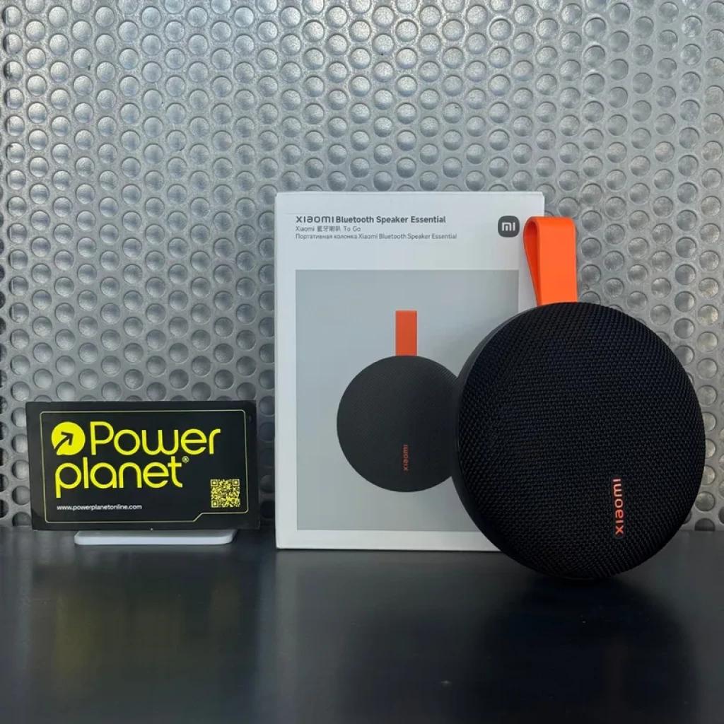 Altavoz portátil Xiaomi Bluetooth Speaker Essential negro, forma redonda con rejilla textil, correa naranja para colgar, caja blanca al fondo y soporte de