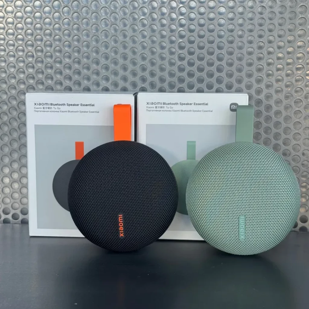 Altavoz portátil Xiaomi Essential en negro, diseño circular con malla textil y correa, compacto. Caja muestra variantes en negro y verde menta. IP66, 5W, BT