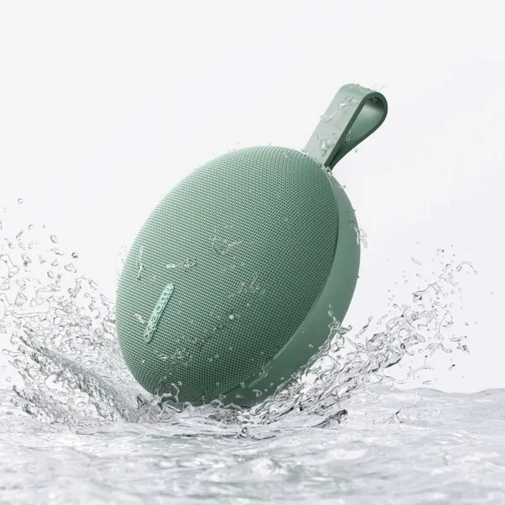 Altavoz portátil verde redondo con rejilla fina, correa de silicona y diseño compacto; resistente al agua con certificación IP66, chapoteando en agua