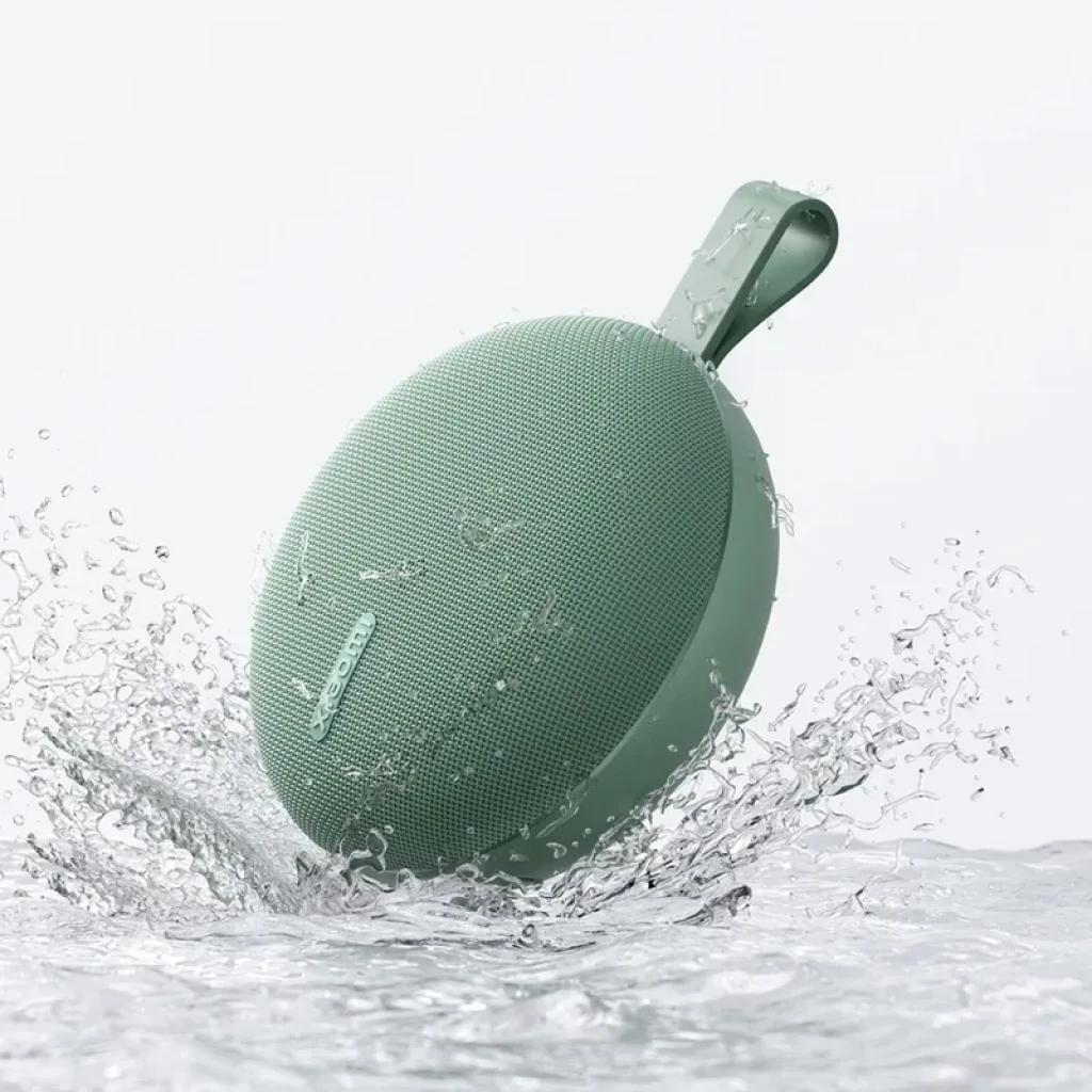 Altavoz portátil verde redondo con rejilla fina, correa de silicona y diseño compacto; resistente al agua con certificación IP66, chapoteando en agua