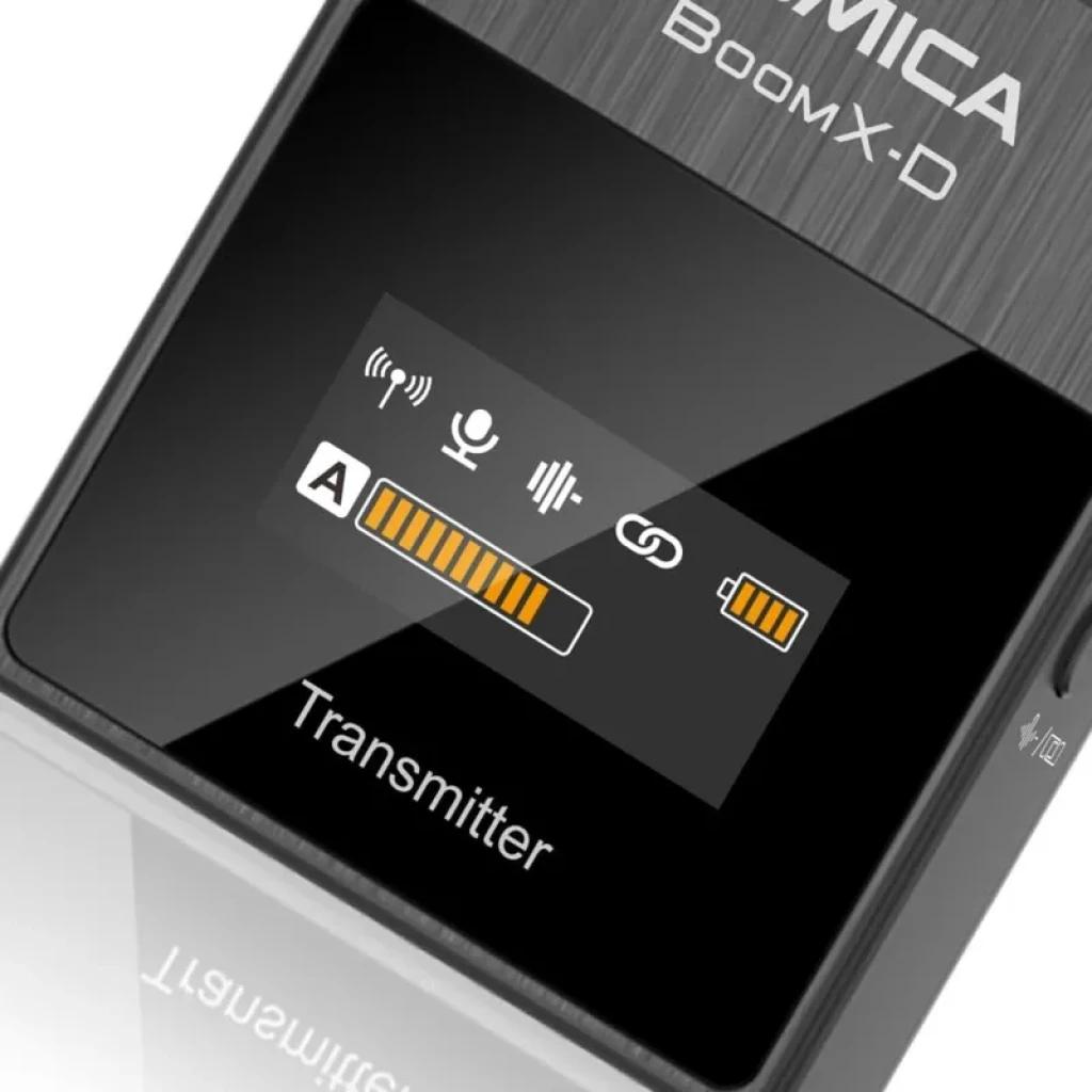 Transmisor Comica BoomX-D D1 negro con pantalla OLED: iconos de audio, enlace y batería, indicadores A y niveles naranjas; acabado metálico cepillado