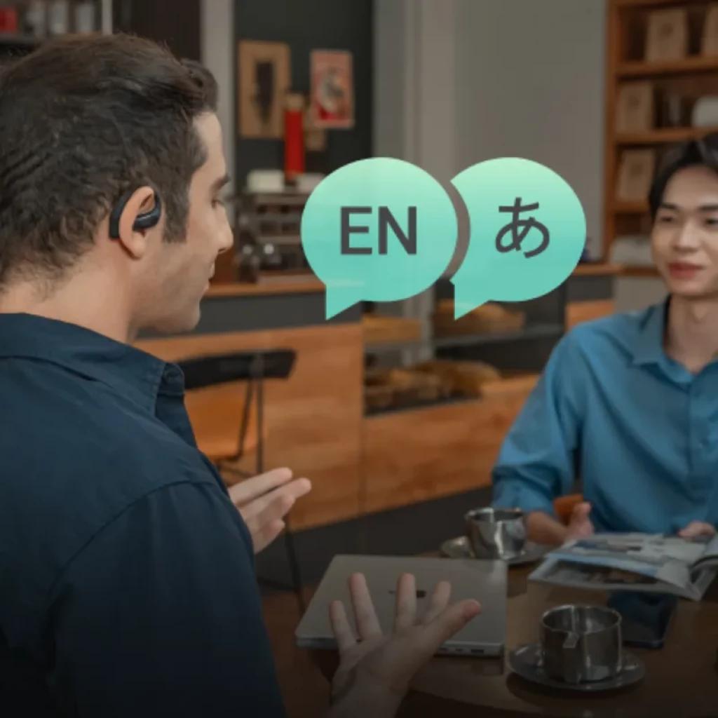 TicNote Pods 4G open-ear negros en uso durante conversación; micrófono lateral discreto, burbujas EN y japonés, ambiente de cafetería, taza y revista en