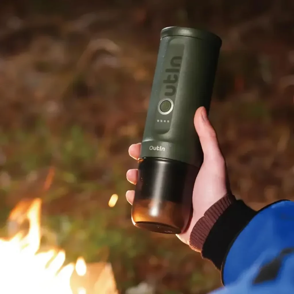 Cafetière électrique portable Outin Nano vert kaki, compacte, corps texturé avec indicateurs LED, base noire, tenue en main près d un feu en plein air