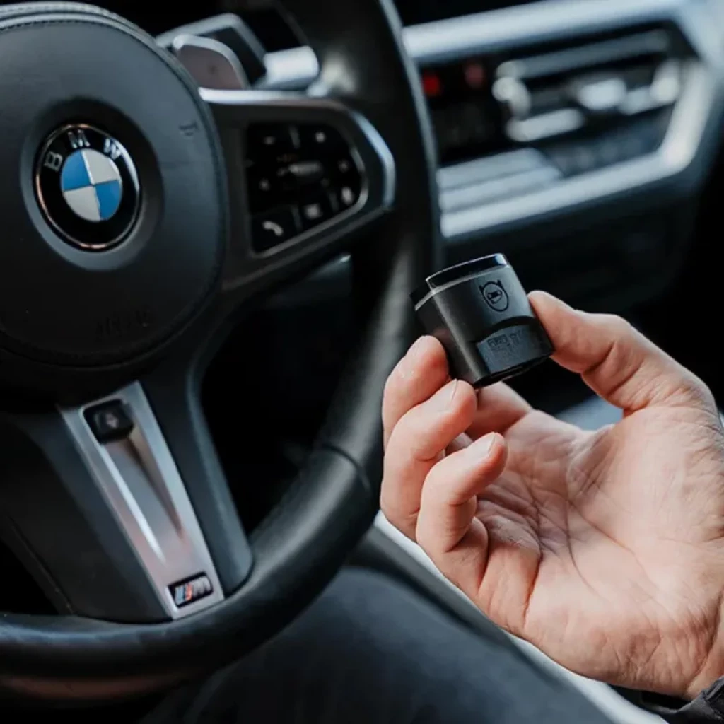 Adaptador OBD compacto negro con logo OBDeleven, diseño cilíndrico y luces indicadoras, sujeto en mano frente a interior de coche moderno con volante