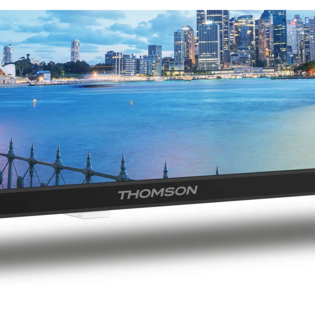 Televisor THOMSON 24 pulgadas con bisel negro delgado, logotipo frontal plateado y pantalla mostrando ciudad costera; ideal para camper con adaptador 12V