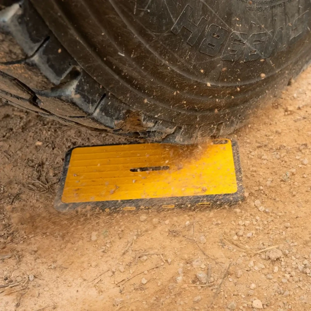 Tableta robusta amarilla bajo la rueda de un vehículo, cubierta con polvo y tierra; carcasa antideslizante negra, diseño resistente y duradero para uso
