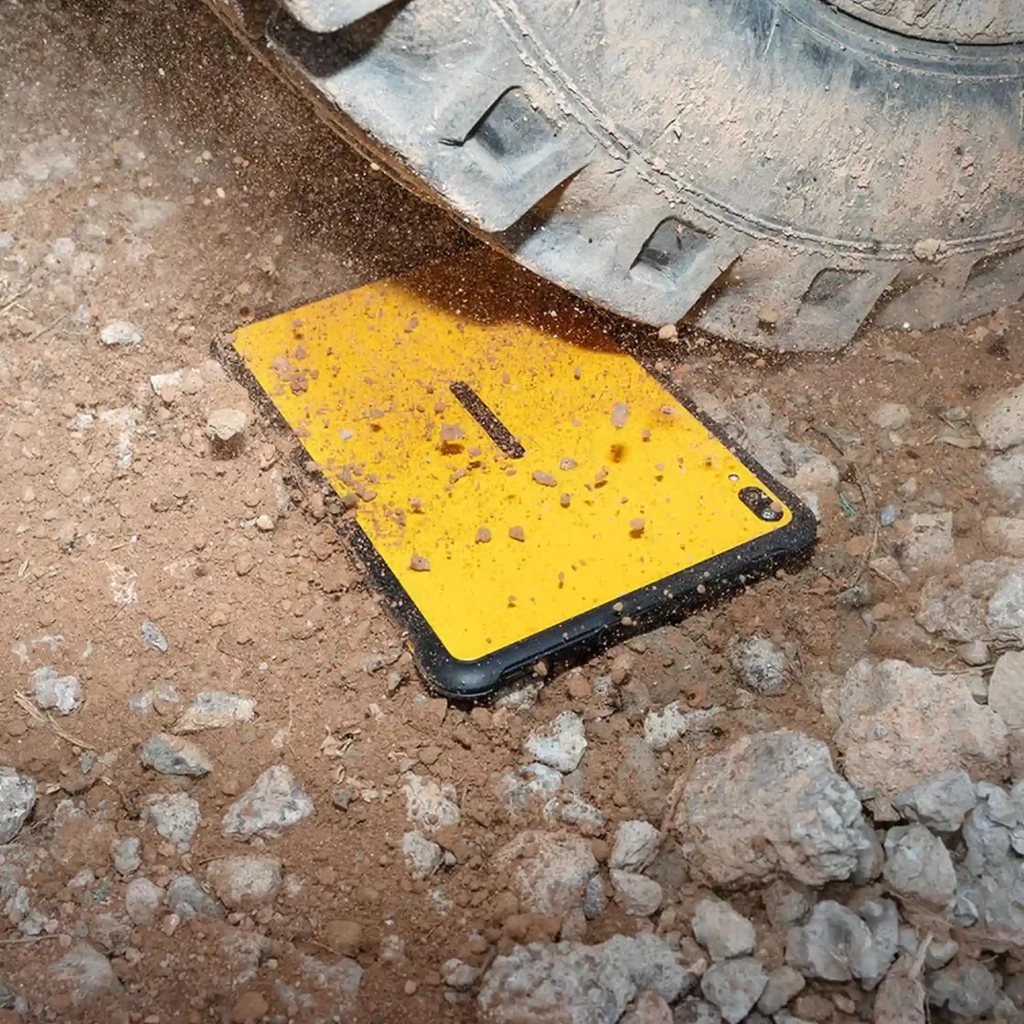 Tableta rugerizada color amarillo y negro bajo una llanta en tierra, cuerpo resistente con marco reforzado y cámara trasera visible en la esquina superior