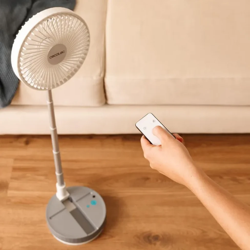 Ventilador de pie plegable con base circular gris, mástil telescópico y rejilla blanca; incluye control remoto y botones azules en la base, diseño compacto