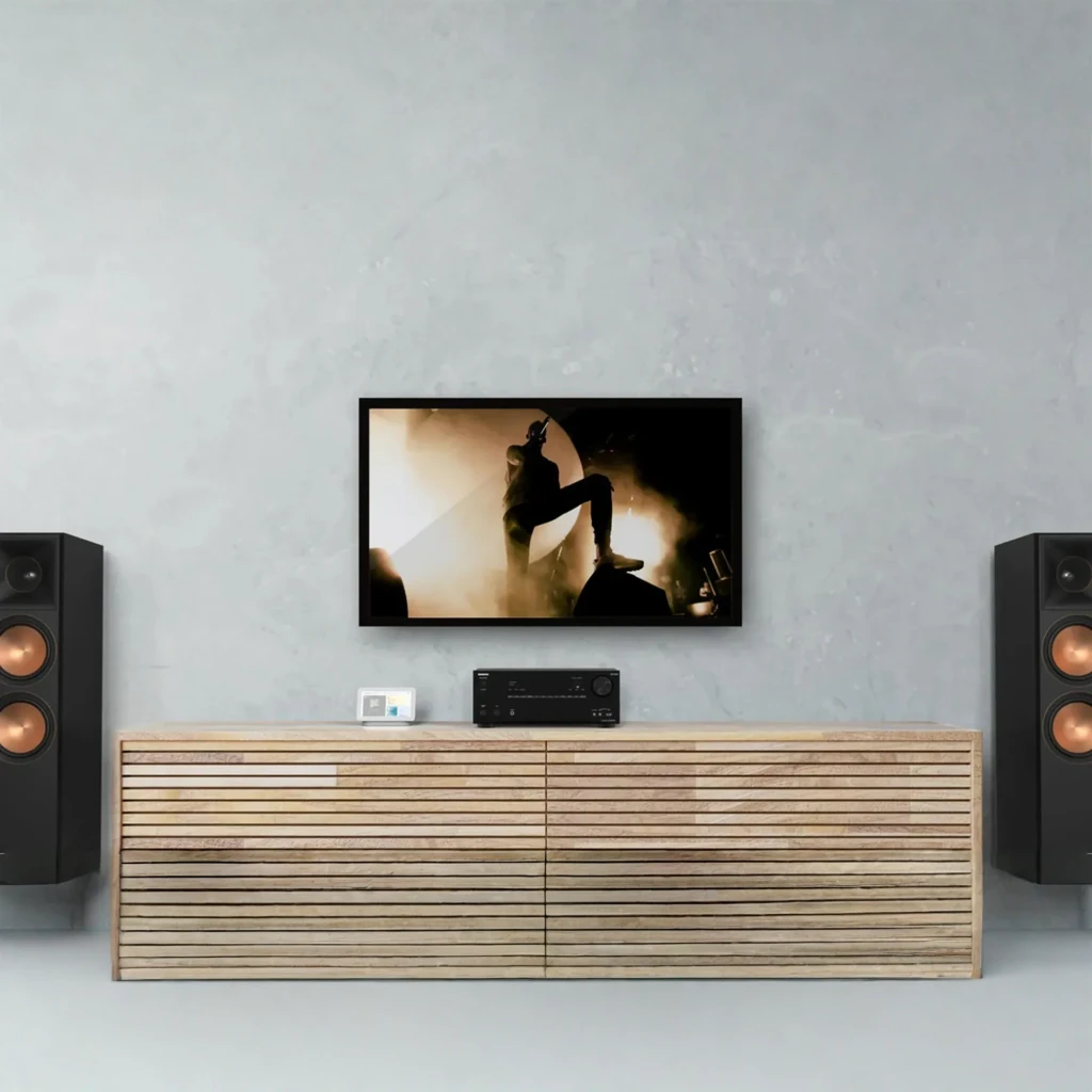 Receptor AV Onkyo TX-NR7100 negro en mueble minimalista, pantalla montada en pared y dos altavoces torre; panel frontal con perilla grande y display iluminado