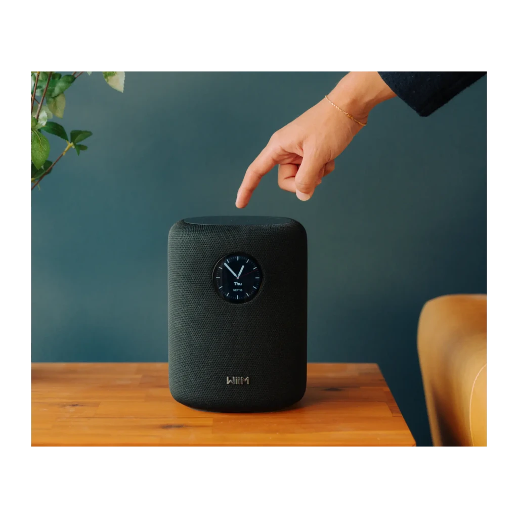 Wiim Sound negro, uso: mano señalando la parte superior, pantalla circular con reloj visible en tela negra sobre mesa de