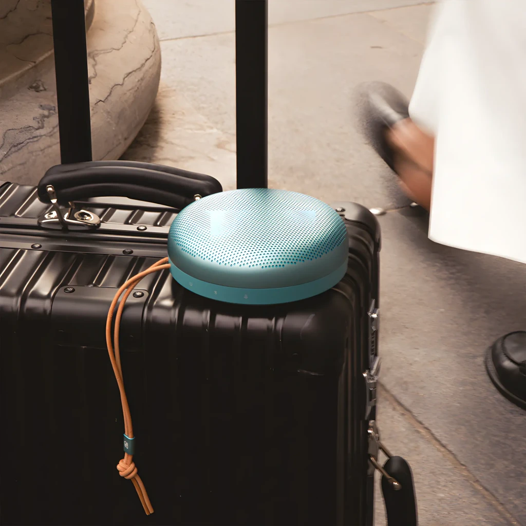 Bang & Olufsen Beosound A1 3ª Gen verde eucalipto, vista lifestyle: altavoz sobre maleta negra con correa de cuero en