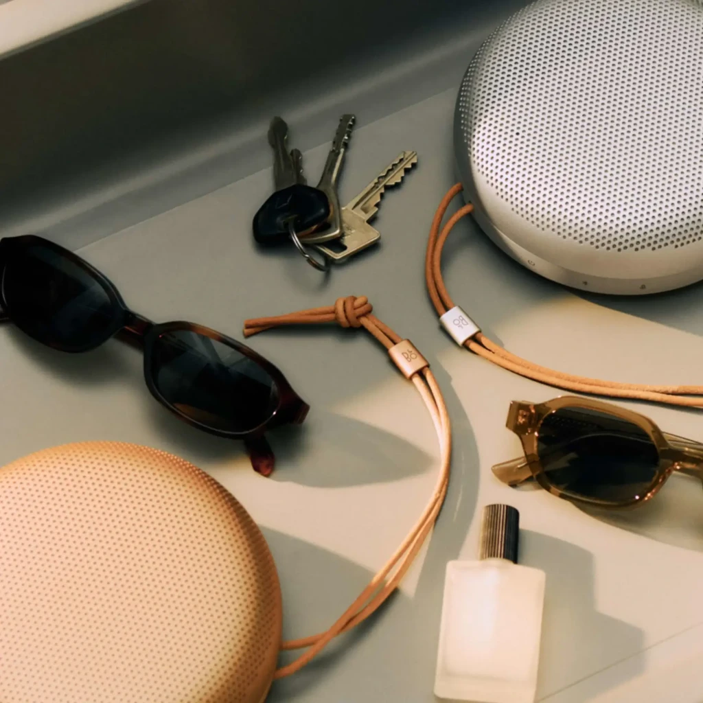 Bang & Olufsen BeoSound A1 3rd Gen Rojo Rústico, lifestyle: dos altavoces con correa de cuero junto a gafas y llaves