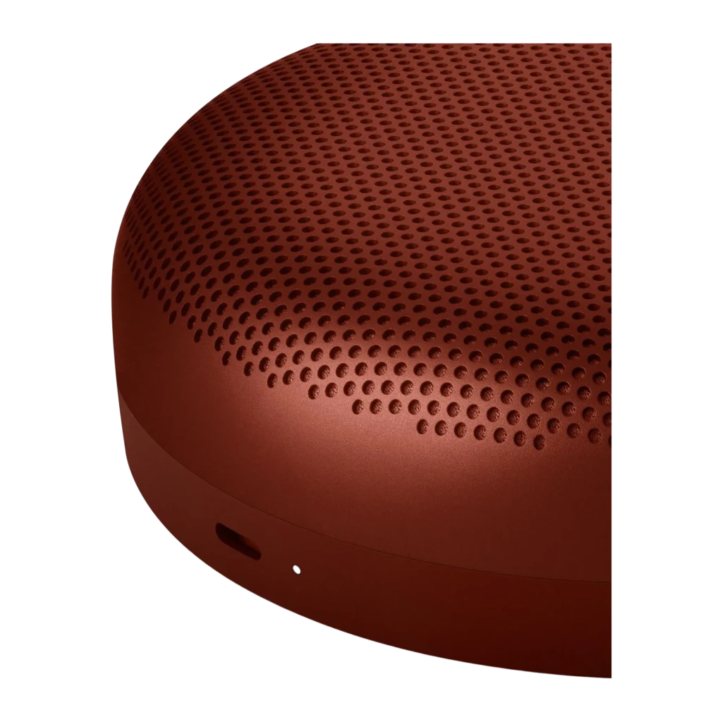Bang & Olufsen BeoSound A1 3rd Gen rojo, detalle lateral: rejilla perforada y botón en la base sobre fondo oscuro