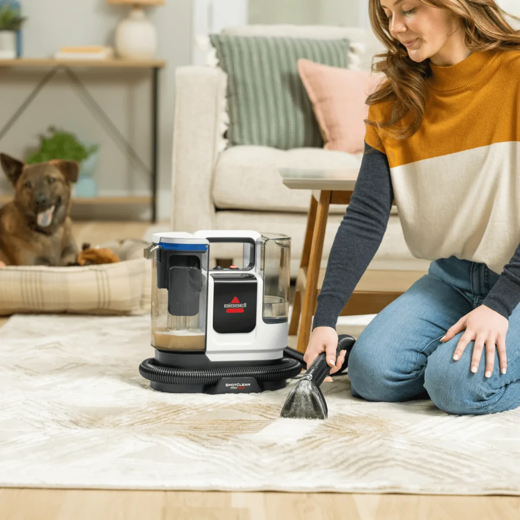 BISSELL SpotClean ProHeat 36988 Rojo Titanio, uso: mujer limpiando mancha en alfombra con accesorio, perro al fondo