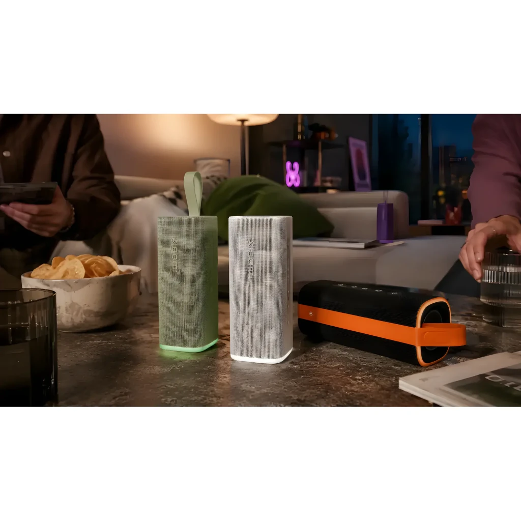 Altavoz portátil verde con forma rectangular, rejilla textil clara, base luminosa, asa de silicona y diseño resistente, junto a variante en gris y funda