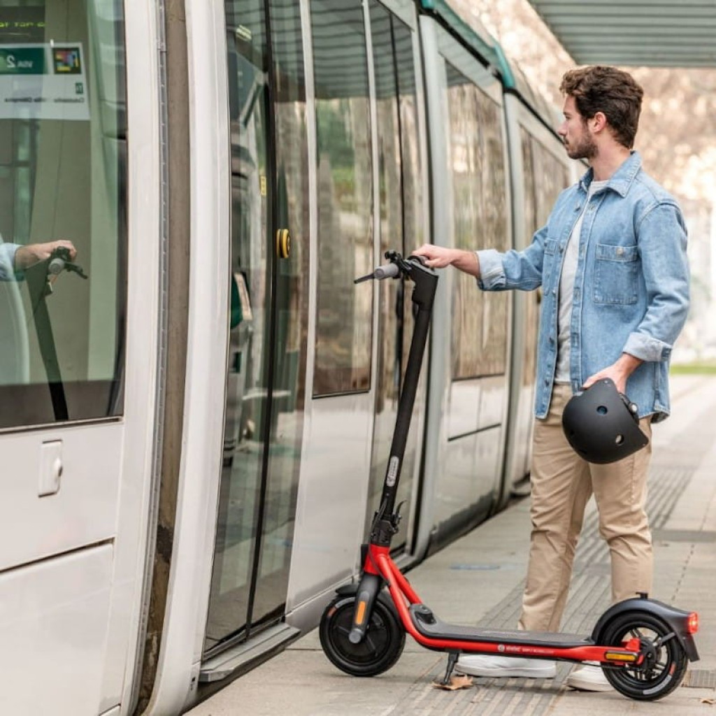 Segway Ninebot KickScooter D18E en color negro y rojo, manillar vertical, ruedas grandes, diseño plegable y ligero; usuario con casco junto a tranvía