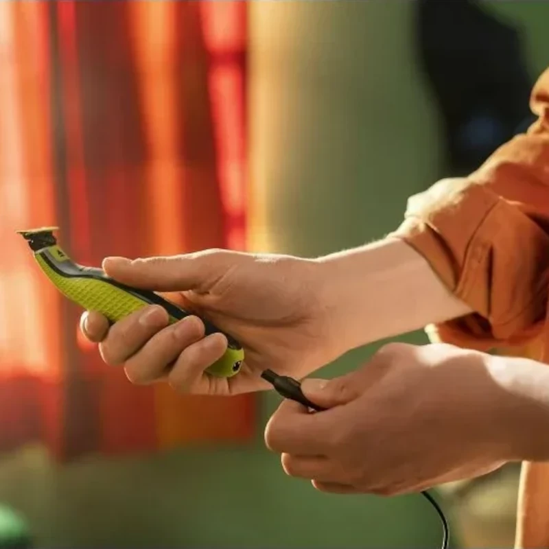 Aparelho Philips OneBlade QP2724/10 em mãos, lâmina verde com protetor e cabo de carregamento conectado, design ergonômico e compacto para barbear e aparar