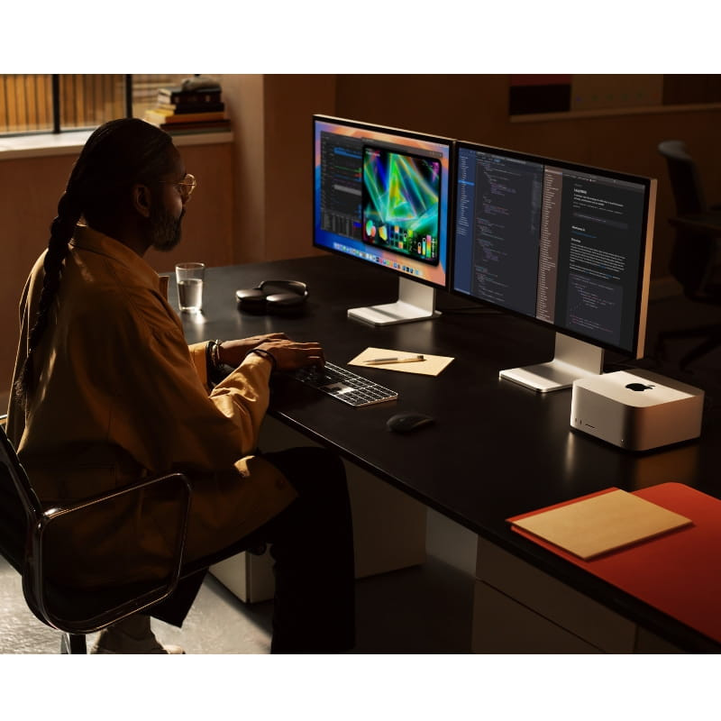 Apple Mac Studio plata compacto sobre escritorio, diseño minimalista con puertos frontales; setup con dos monitores, teclado y ratón en ambiente profesional