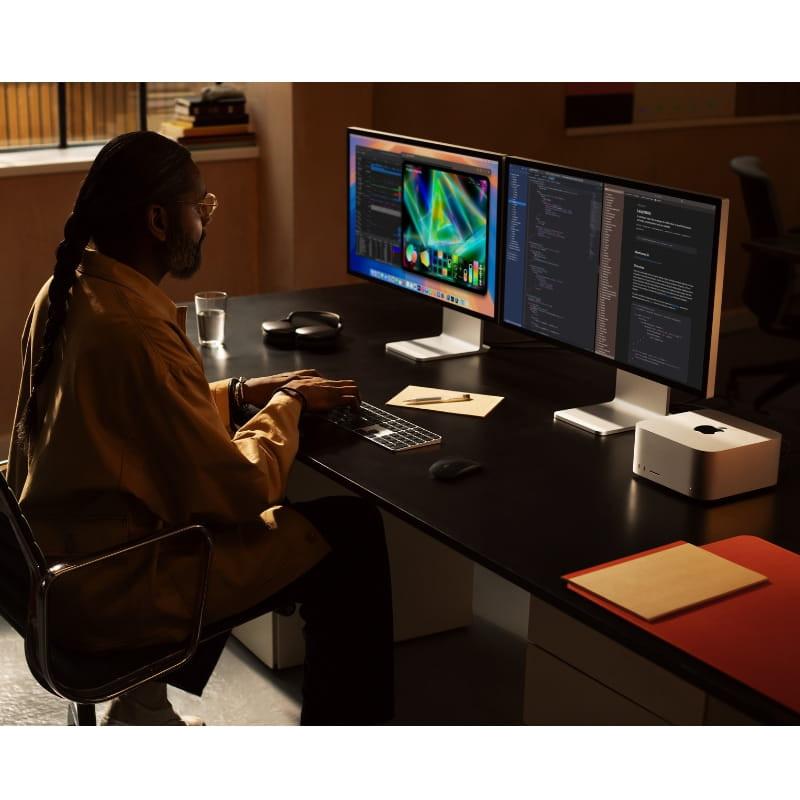 Apple Mac Studio plata compacto sobre escritorio, diseño minimalista con puertos frontales; setup con dos monitores, teclado y ratón en ambiente profesional