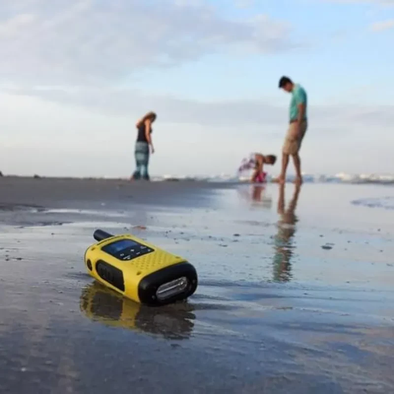 Walkie talkie amarillo y negro Motorola Talkabout T92 H2O sobre arena húmeda; diseño robusto, resistente al agua, pantalla y botones grandes, clip metálico