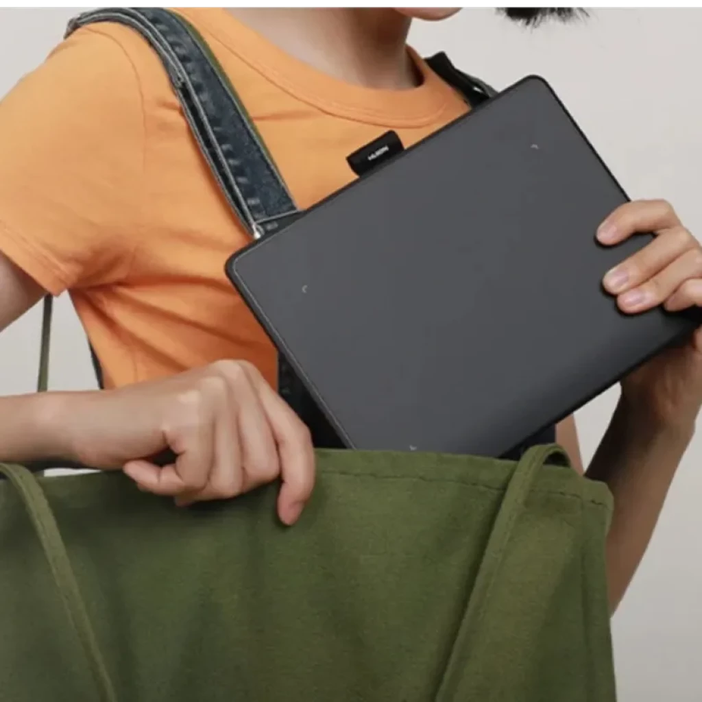 Tablette graphique compacte gris anthracite rangée dans un sac vert, bords arrondis, surface lisse, format portable; main tenant la tablette par le côté