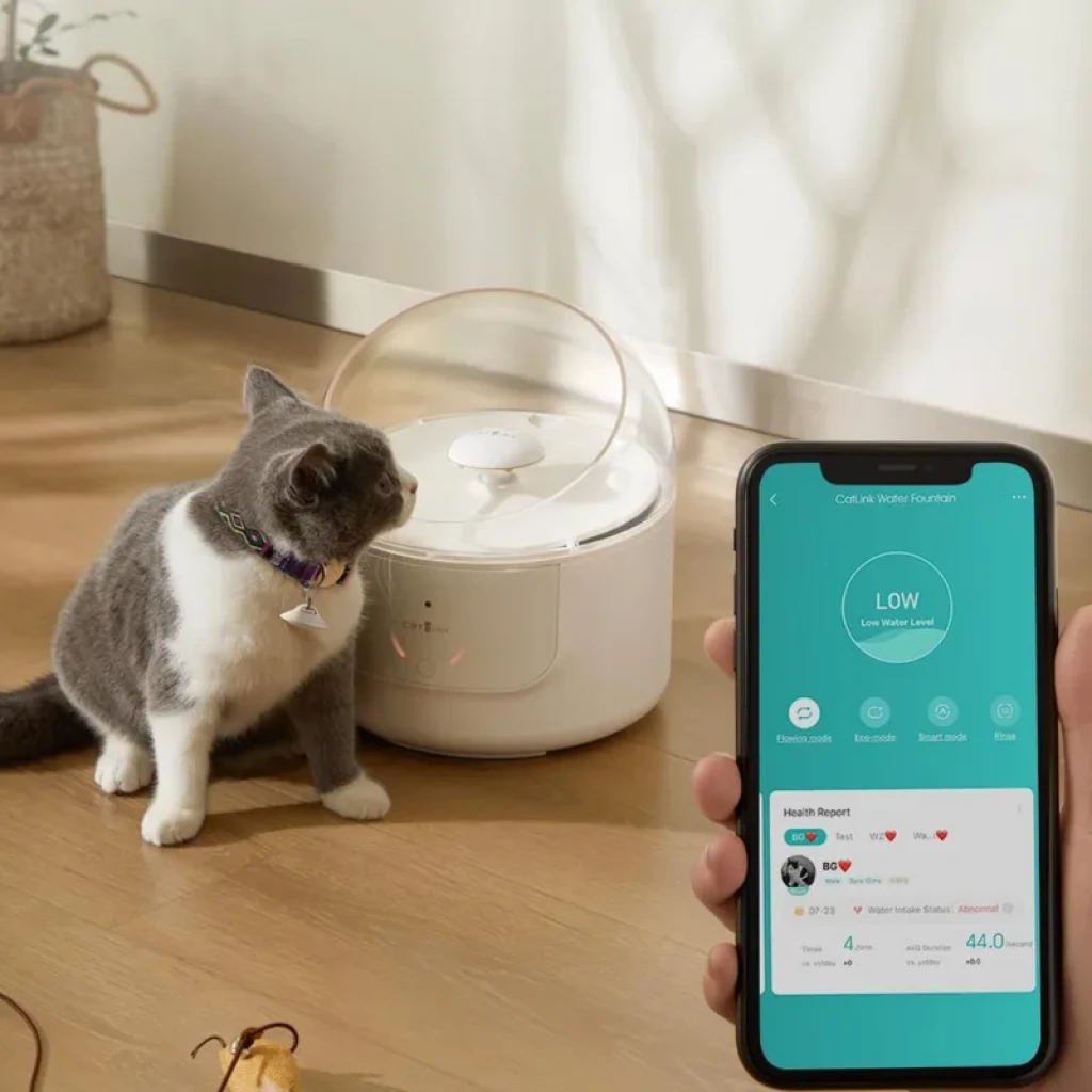 Fuente inteligente CATLINK blanca con cúpula transparente y luz LED, gato gris observando, app móvil mostrando nivel de agua y datos de salud en pantalla