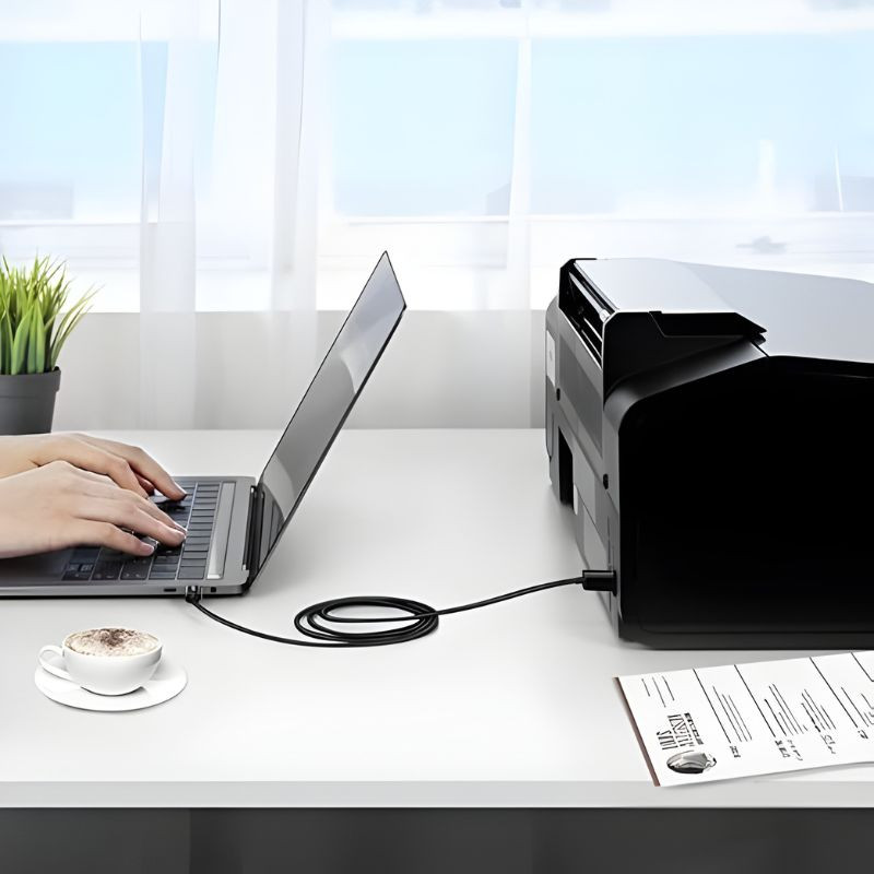 Cable Ugreen USB-C a USB-B de 2 m conectando laptop a impresora; diseño negro, conector USB-B cuadrado y USB-C delgado, mesa blanca y cable enrollado